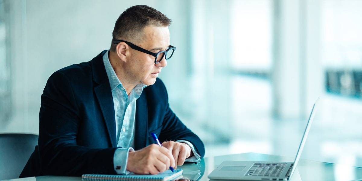 Male with blue suite on sitting at table taking notes from his laptop.