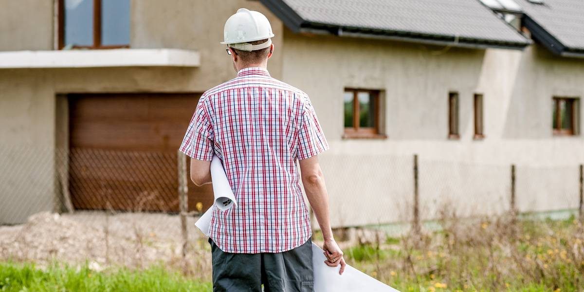 Male construction worker wearing white helmet walking towards a brown house.